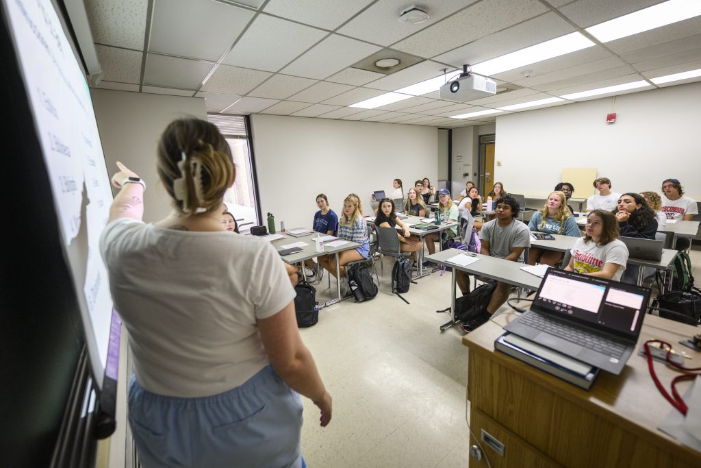 Photo of an instructor standing in front of a class of students, speaking to them while pointing to a slide on a screen.