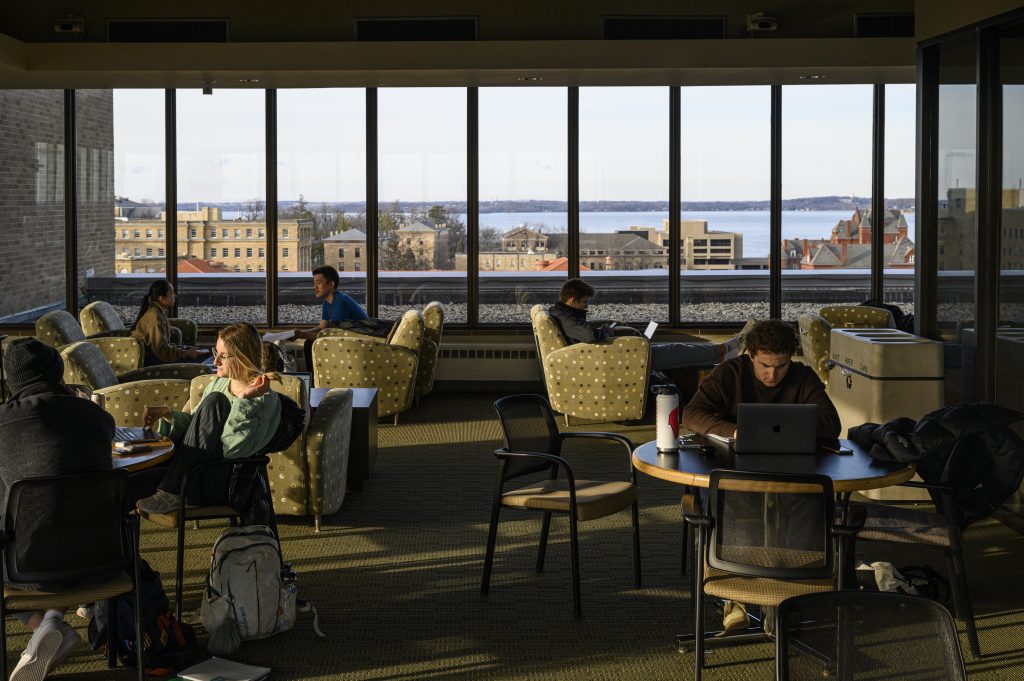 Photo of students studying and chatting at round tables in the lounge of a high-rise building with windows looking out over Lake Mendota.
