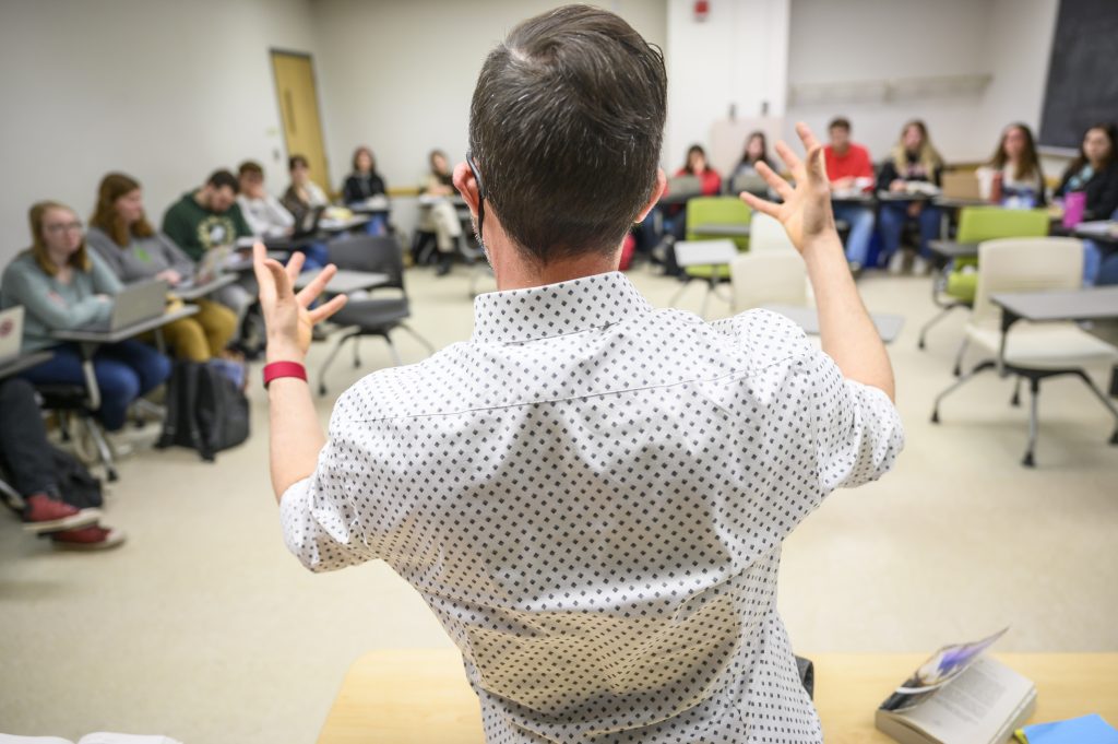 Photo of an instructor with his back to the photographer, facing a small classroom of students in individual desks and speaking while gesturing with his hands.