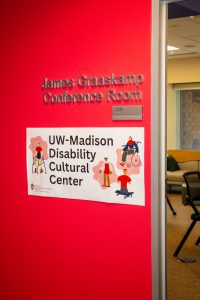 Red wall and open door to the DCC with metal sign that reads “James Graaskamp Conference Room" and room number “2096,” along with a banner that reads “UW-Madison Disability Cultural Center.”