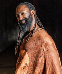 Antoine Hunter is a Black and Indigenous person who has dark brown skin, full beard, and long black locs. Antoine is wearing a brown shawl and smiling.
