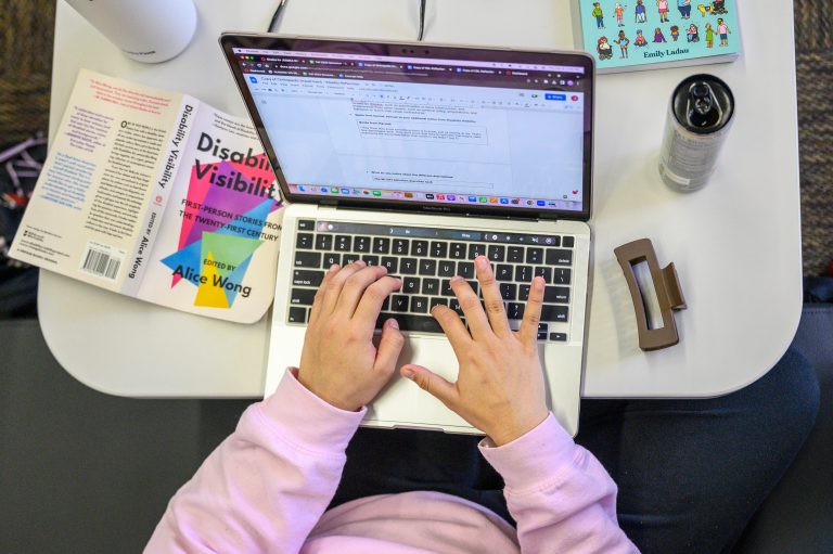 A student sitting at a computer with an open book called Disability Visibility by Alice Wong.